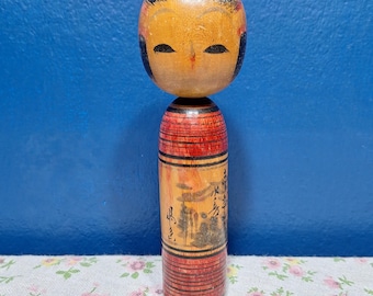 May include: A traditional Japanese Kokeshi doll with a round head and cylindrical body. The doll's head is painted with a yellow face, black eyes, and black hair. The body is red and orange with a painted landscape scene and Japanese characters.
