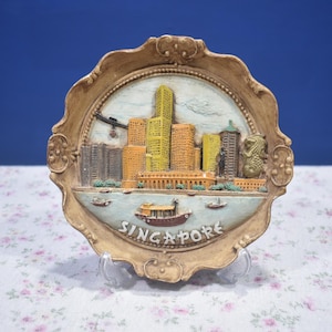 Pode incluir: Um prato decorativo e ornamentado com um relevo do horizonte de Singapura. O prato tem uma moldura castanha clara e uma imagem central com edifícios, barcos e o texto "SINGAPORE". O prato está sobre um suporte transparente.