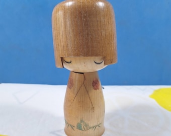 May include: A wooden Kokeshi doll with a rounded head and a cylindrical body. The doll features painted closed eyes, a small bow, and floral and leaf designs. The wood has a natural, light brown color.