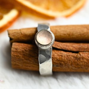 Op de afbeelding: Een zilveren ring met een gehamerde textuur en een rond, licht verhoogd, koperkleurig middelste deel. De ring ligt op twee kaneelstokjes.