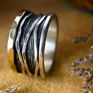 May include: A wide silver and gold band ring with a textured, hammered finish. The ring is made of multiple bands of metal that are woven together.