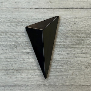 May include: A solid black, three-dimensional triangular prism object with sharp, clean edges and a smooth, reflective surface. The geometric form tapers to a single point at the bottom, showcasing a uniform dark finish across all visible sides.