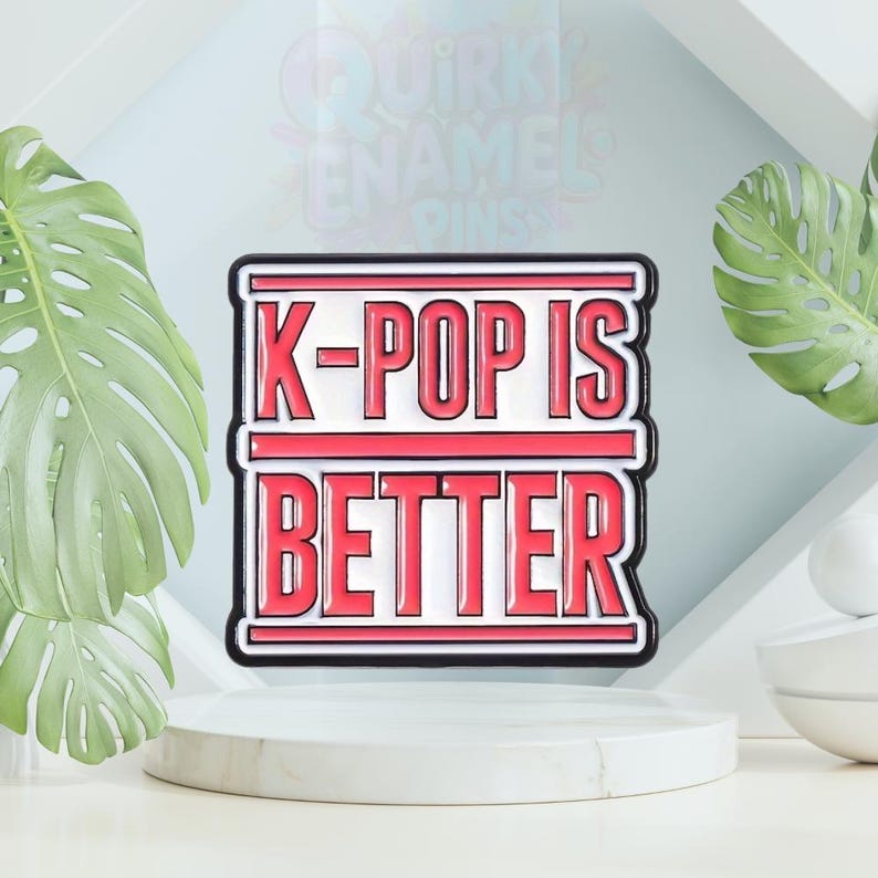 以下が含まれることがあります： 赤と白で「K-POP IS BETTER」の文字が入った長方形のエナメルピン。ピンは黒い縁取りがあり、背景に緑の葉がある白い台座に飾られています。