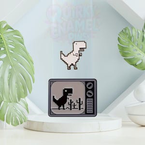 Könnte beinhalten: Zwei Emaille-Anstecknadeln mit einem pixeligen Dinosaurier-Design. Eine ist ein weißer Dinosaurier, die andere ein schwarzer Dinosaurier in einem grauen Fernseher. Die Anstecknadeln werden auf einem weißen Sockel mit grünen Blättern präsentiert. Der Text "Quirky Enamel Pins" ist sichtbar.