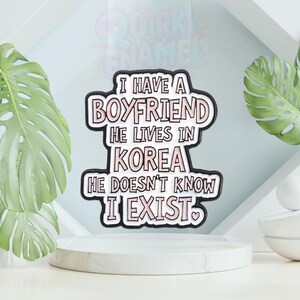 以下が含まれることがあります： 「I HAVE A BOYFRIEND HE LIVES IN KOREA HE DOESN'T KNOW I EXIST」のテキストが入った白とピンクのエナメルピン。ピンには黒い縁取りとハートのシンボルが付いています。ピンは白い台座にあります。