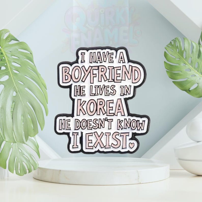 以下が含まれることがあります： 「I HAVE A BOYFRIEND HE LIVES IN KOREA HE DOESN'T KNOW I EXIST」のテキストが入った白とピンクのエナメルピン。ピンには黒い縁取りとハートのシンボルが付いています。ピンは白い台座にあります。