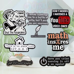 May include: A collection of eight enamel pins with humorous sayings. The pins feature black and white illustrations and colourful text. The sayings include "I baked you some, Shut the Fucupcakes", "I would challenge you to a battle of wits, but I see you are unarmed. -William Shakespeare", "Don't worry I watched a YouTube video once", "There is no place like 127.0.0.1", "Math inspires me", "Just one more chapter", and "Have you tried turning it off & on again?"