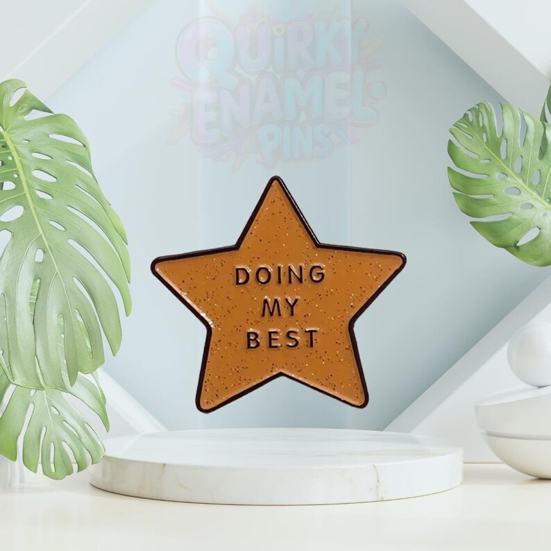 May include: A star-shaped enamel pin in shades of brown with the words "DOING MY BEST" in a textured font. The pin is set against a light blue backdrop with green foliage and a white pedestal.