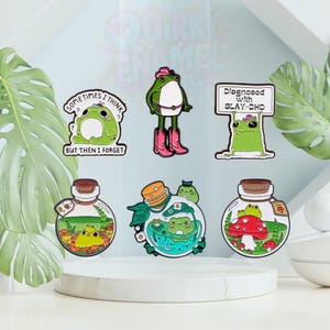 May include: A selection of enamel pins with quirky frog illustrations. One pin depicts a frog in a cowboy hat and boots. Other pins show frogs in glass jars and with the text "Sometimes I think but then I forget" and "Diagnosed with SLAY-DHD."