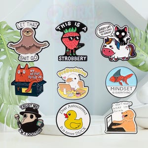 Op de afbeelding: Een verzameling kleurrijke emaille pins met diverse ontwerpen en tekst. De pins tonen een luiaard, aardbei, kat, eenhoorn, kat in een doos, eend, vis, pestdokter, eend en hond. Teksten: "Let that shit go", "This is a Strobbery", "Giddy up! There's chaos to spread!", "No desire to fit in", "ProDucktive", "Mindset is everything", "Trust me I'm a doctor", "Dear autocorrect it is never duck", en "I don't know what I'm doing".