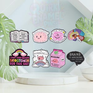 May include: A selection of quirky enamel pins with different designs. The pins feature a brain, an open book, a rainbow, and the text "I don't have enough serotonin for this shit". Other pins have phrases such as "Little Miss Procrastinator" and "Brains are awesome".