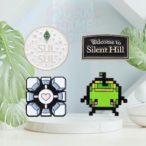 Könnte beinhalten: Vier Emaille-Anstecknadeln werden gezeigt. Eine ist ein weißer Kreis mit einem grünen Edelstein und dem Wort "SUL". Eine andere ist ein schwarzes Schild mit der Aufschrift "Welcome to Silent Hill". Eine dritte ist ein Würfel mit einem Herzen. Die letzte ist ein grüner Pixel-Charakter.