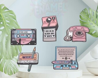 May include: A collection of enamel pins featuring retro designs. Includes a cassette tape with "FINE LINE" text, a pink camera with "I SEE US IN BLACK AND WHITE" text, a pink rotary phone, a cassette tape with a pencil, and a record player.