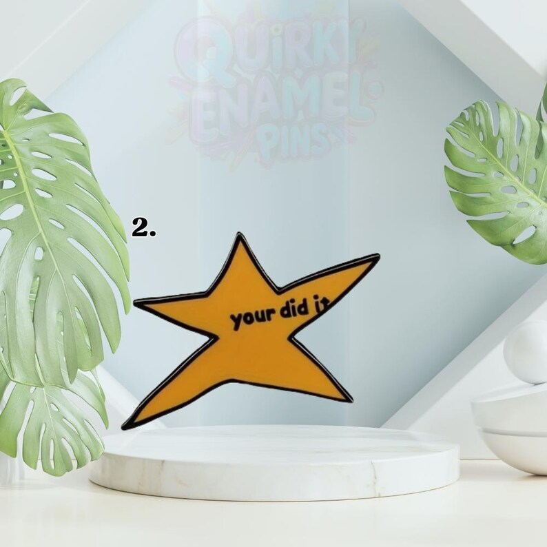May include: A gold star-shaped enamel pin with a black outline and the words "your did it" in black. The pin is displayed on a white pedestal with a light blue background and green leaves.