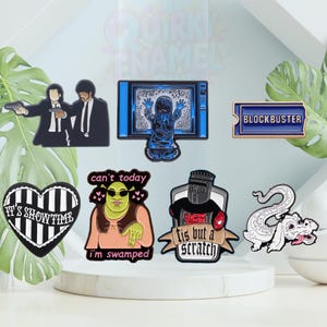 May include: A selection of enamel pins with pop culture themes. Designs include characters from Pulp Fiction, a TV with a ghostly figure, a Blockbuster logo, a heart with "It's Showtime", Shrek, a knight, and a dragon.