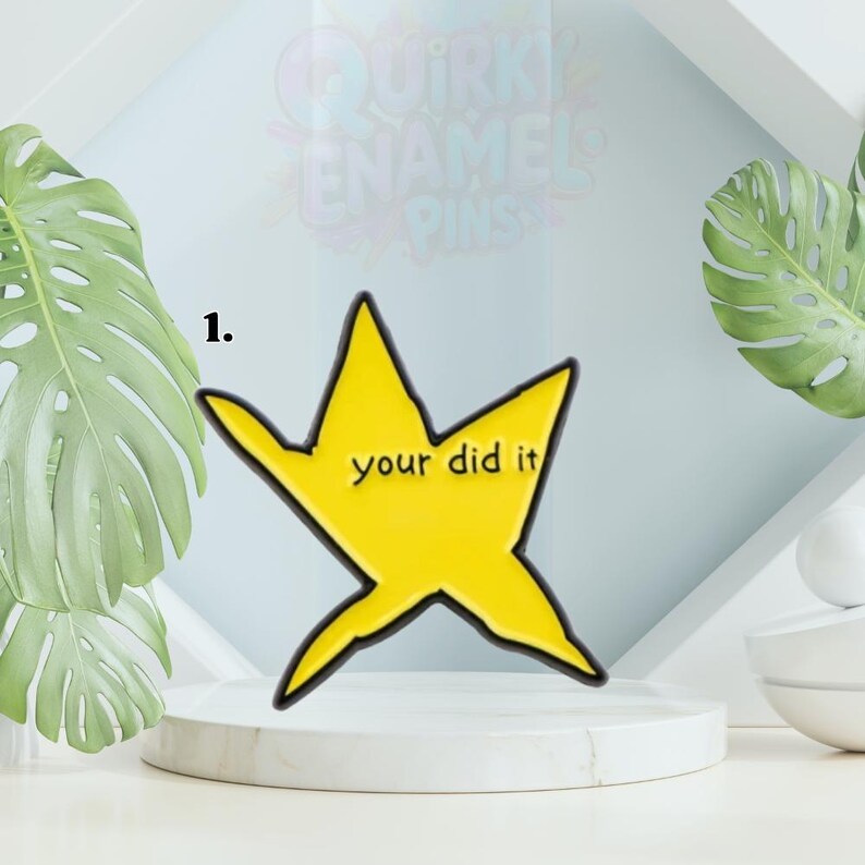 May include: A yellow star-shaped enamel pin with a black outline and the words "your did it" in a black font. The pin is displayed on a white pedestal with a light blue background and green leaves.