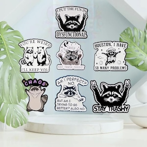 Op de afbeelding: Een verzameling stickers met wasbeer-thema's en verschillende zinnen. De stickers tonen wasberen in verschillende poses, met teksten als "Dysfunctional", "Stay Trashy" en "Chaos". Het kleurenpalet omvat zwart, wit en bruin.