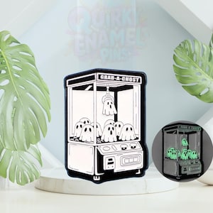 Pode incluir: Pin de esmalte preto e branco com uma máquina de garra com o texto "GRAB-A-CHOST". A máquina está cheia de figuras de fantasmas. O pin tem uma função que brilha no escuro. O pin é exibido em uma superfície branca com folhas verdes no fundo.
