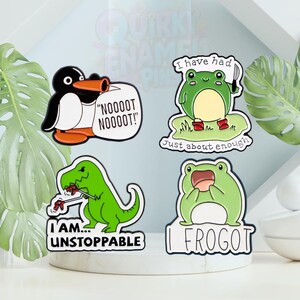 May include: Four enamel pins with quirky designs. A penguin with a speech bubble saying "NOOOOT NOOOOT!". A frog holding a knife with the text "I have had just about enough". A green dinosaur with "I AM... UNSTOPPABLE". A frog with "I FROGOT".