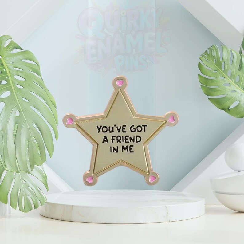 May include: A gold star-shaped enamel pin with the text "YOU'VE GOT A FRIEND IN ME". The pin has pink heart-shaped accents on each point. The background includes a white pedestal, green leaves, and a light blue backdrop.
