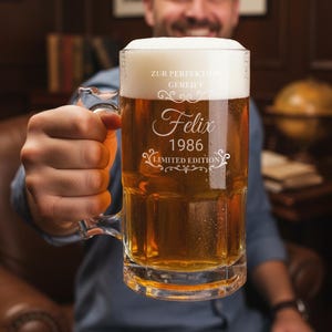 Op de afbeelding: Een heldere glazen bierpul gevuld met bier en schuim, vastgehouden door een persoon. De pul heeft een handvat en is gegraveerd met de naam "Felix", het jaar "1986" en de woorden "Limited Edition".