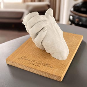 May include: A white plaster hand casting of two hands holding each other, displayed on a wooden base with the inscription "Susan & William 05.09.2025".