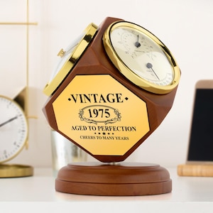 Könnte beinhalten: Eine Holzuhr mit goldenem Zifferblatt und goldener Einfassung. Die Uhr hat eine goldene Plakette mit dem Text "Vintage 1975 Aged to Perfection Cheers to Many Years".