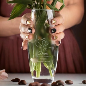 Può includere: Un vaso di vetro trasparente con un design inciso di due cuori e il testo "Mr & Mrs Davies 40 Years Married 10.22.2021 Love Jack & Melanie". Il vaso è pieno di fiori rosa e bianchi e acqua.