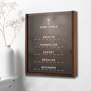 Puede incluir: Una escala de vino de madera marr&oacute;n con un frente de vidrio. La escala tiene un icono de copa de vino blanco y el texto "Wine Scale" en la parte superior. Debajo del texto hay cinco niveles: Kristin, Sommelier, Experto, Regular y Principiante.