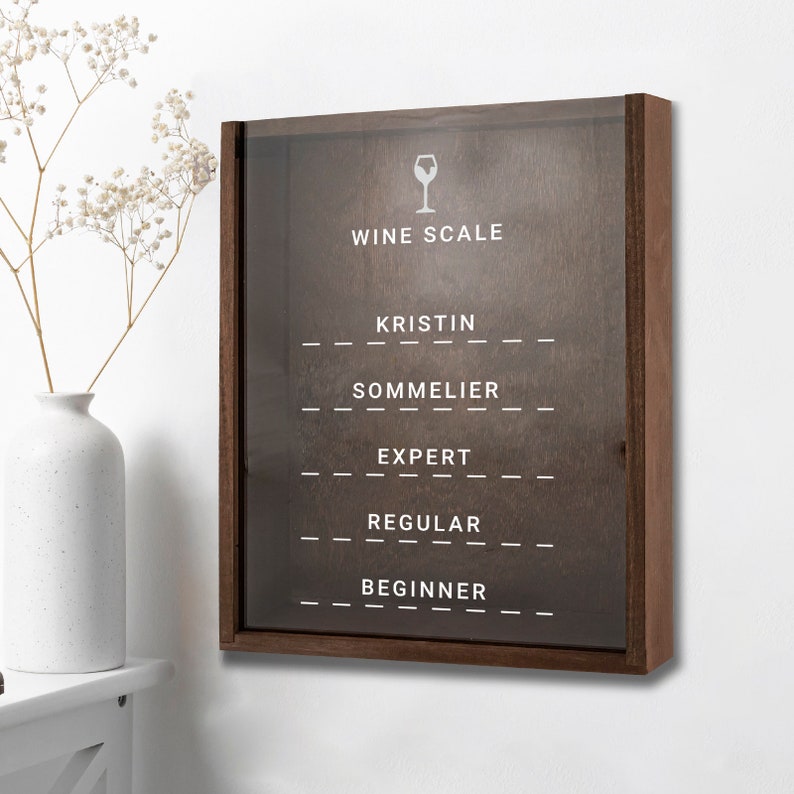 Puede incluir: Una escala de vino de madera marr&oacute;n con un frente de vidrio. La escala tiene un icono de copa de vino blanco y el texto "Wine Scale" en la parte superior. Debajo del texto hay cinco niveles: Kristin, Sommelier, Experto, Regular y Principiante.