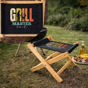 Puede incluir: Un taburete plegable de madera con un asiento negro con las palabras "GRILL MASTER" en una fuente retro. El diseño incluye una silueta de una persona asando a la parrilla. El taburete está instalado al aire libre cerca de una parrilla.
