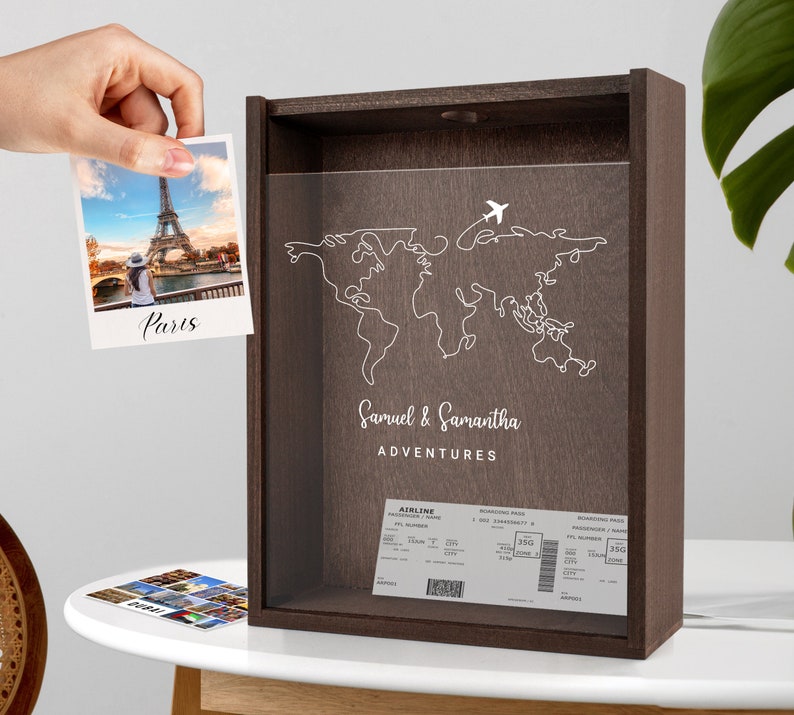 May include: A wooden shadow box with a world map outline and the text "Samuel & Samantha Adventures". The box has a slot for inserting photos and a clear acrylic front. A photo of the Eiffel Tower in Paris is inserted in the slot.