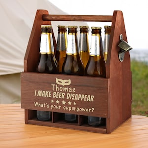 Peut inclure: Un porte-bière en bois brun pour six bouteilles avec un ouvre-bouteille intégré. Le porte-bière a une poignée et est personnalisé avec le texte "Thomas I MAKE BEER DISAPPEAR What's your superpower?" et un graphique de masque doré.