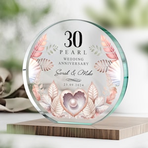 Può includere: Targa rotonda in vetro trasparente per il 30° anniversario di matrimonio. La targa presenta la scritta "30 PEARL WEDDING ANNIVERSARY", nomi e una data. Il design include una perla a forma di cuore e foglie decorative.