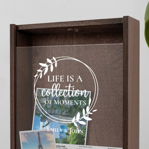 Peut inclure: Une bo&icirc;te &agrave; ombre en bois avec un devant en verre transparent. La bo&icirc;te porte une inscription blanche qui dit "Life is a collection of moments" avec un motif floral. La bo&icirc;te est remplie de photos et de souvenirs. L'inscription est sign&eacute;e "Emily & John".