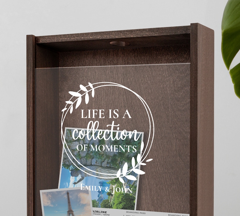 Peut inclure: Une bo&icirc;te &agrave; ombre en bois avec un devant en verre transparent. La bo&icirc;te porte une inscription blanche qui dit "Life is a collection of moments" avec un motif floral. La bo&icirc;te est remplie de photos et de souvenirs. L'inscription est sign&eacute;e "Emily & John".