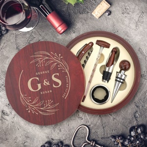 Könnte beinhalten: Eine Holzkiste mit roter Oberfläche und einem Deckel, der mit einem goldenen Blumenmuster und dem Text "George & Susan Est. 7.1.2023" verziert ist. Die Kiste enthält einen Flaschenöffner, einen Flaschenverschluss, ein Thermometer und einen Foliencutter.