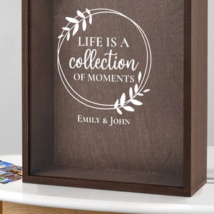 Peut inclure: Une bo&icirc;te &agrave; ombre en bois brun avec un devant en verre. La bo&icirc;te porte une inscription blanche qui dit "Life is a collection of moments" avec une couronne florale autour du texte. L'inscription comprend &eacute;galement les noms "Emily & John".