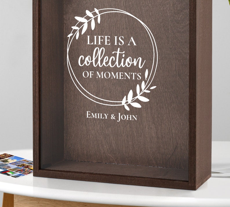 Peut inclure: Une bo&icirc;te &agrave; ombre en bois brun avec un devant en verre. La bo&icirc;te porte une inscription blanche qui dit "Life is a collection of moments" avec une couronne florale autour du texte. L'inscription comprend &eacute;galement les noms "Emily & John".