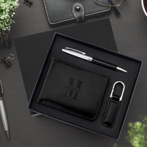 Può includere: Un set regalo nero con portafoglio, penna e portachiavi, tutti ordinatamente disposti in una scatola nera. Il portafoglio e il portachiavi sono impressi con il nome "MICHAEL" e un monogramma. La penna ha un fusto argentato e dettagli neri.