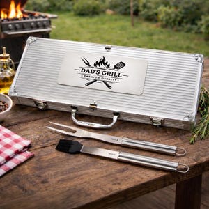 Può includere: Un kit di attrezzi per grigliate argentato con la scritta "DAD'S GRILL - PREMIUM QUALITY" sul coperchio. Il kit include una forchetta e un pennello per spennellare con manici in acciaio inossidabile. Gli attrezzi sono su un tavolo di legno.