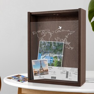 May include: A brown wooden shadow box with a clear acrylic front. Inside the box is a white outline of a world map with a small airplane icon. There are two photos and a boarding pass inside the box. The photos are of the Eiffel Tower and a landscape. The boarding pass is for a flight from Paris to London. The text on the box reads "Samuel & Samantha's Adventures".