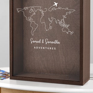 May include: A brown wooden shadow box with a clear glass front. The box features a white outline of a world map with an airplane above it. The text "Samuel & Samantha ADVENTURES" is printed below the map.