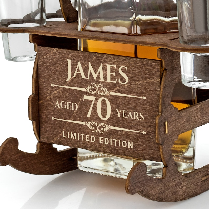 May include: Wooden liquor dispenser shaped like a sleigh with a bottle and four shot glasses. The dispenser is personalized with the name "JAMES" and the text "AGED 70 YEARS LIMITED EDITION".
