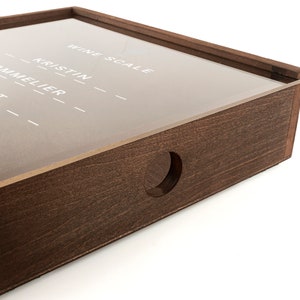 Puede incluir: Una caja de madera con una tapa de acr&iacute;lico transparente. La tapa tiene las palabras "Wine Scale" y "Kristin" escritas en ella. La caja tiene un agujero redondo cortado en el lateral.