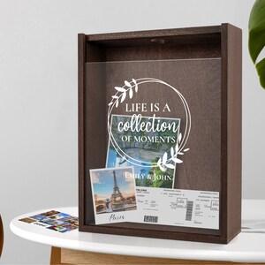 Peut inclure: Une bo&icirc;te en bois brun avec une face en verre. La bo&icirc;te porte une inscription blanche qui dit "Life is a collection of moments" avec un motif floral. La bo&icirc;te contient une photo de la tour Eiffel et deux cartes d'embarquement pour avion. La photo est intitul&eacute;e "Paris" et les cartes d'embarquement sont pour Emily & John.