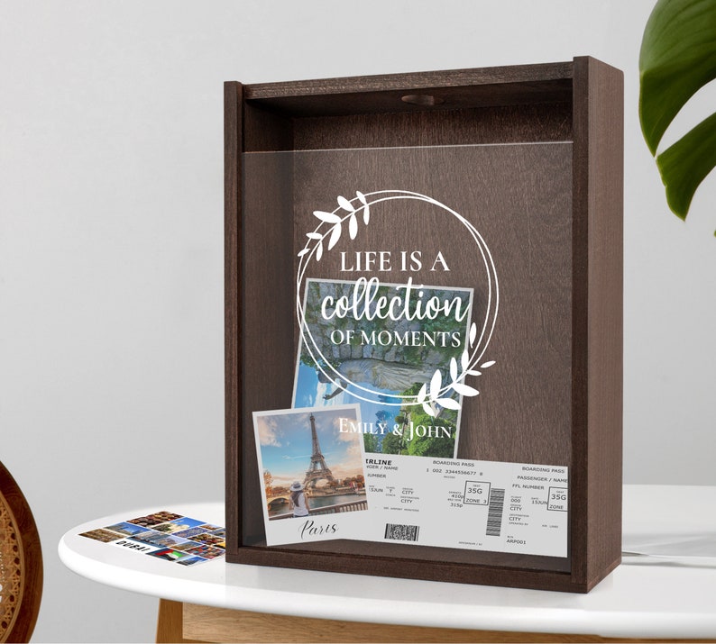 Peut inclure: Une bo&icirc;te en bois brun avec une face en verre. La bo&icirc;te porte une inscription blanche qui dit "Life is a collection of moments" avec un motif floral. La bo&icirc;te contient une photo de la tour Eiffel et deux cartes d'embarquement pour avion. La photo est intitul&eacute;e "Paris" et les cartes d'embarquement sont pour Emily & John.