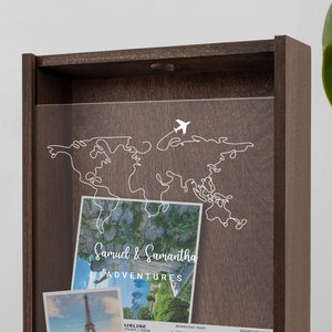May include: A wooden shadow box with a clear acrylic front. The box has a white outline of a world map with a small airplane flying over it. The text "Samuel & Samantha Adventures" is printed on a white background.