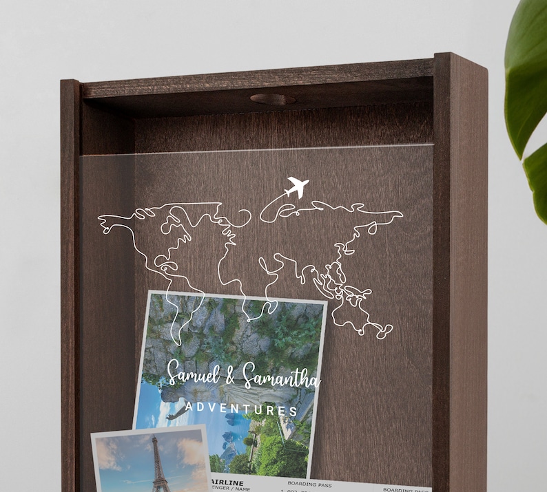 May include: A wooden shadow box with a clear acrylic front. The box has a white outline of a world map with a small airplane flying over it. The text "Samuel & Samantha Adventures" is printed on a white background.