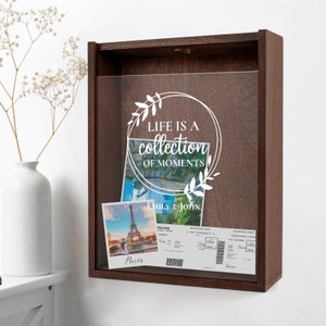 Peut inclure: Une bo&icirc;te &agrave; ombre en bois brun avec un devant en verre transparent. La bo&icirc;te contient deux photos et une carte d'embarquement. Les photos sont de la tour Eiffel &agrave; Paris. La carte d'embarquement est pour un vol vers Paris. La bo&icirc;te a un autocollant en vinyle blanc avec le texte "Life is a collection of moments" et un motif floral.